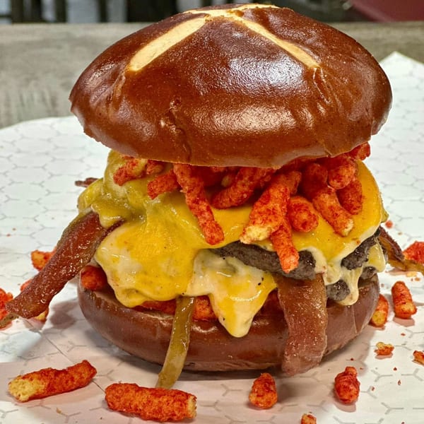 A Market Street Burgers creation: a pretzel bun burger with melted cheese, bacon, and spicy crunchy snacks.