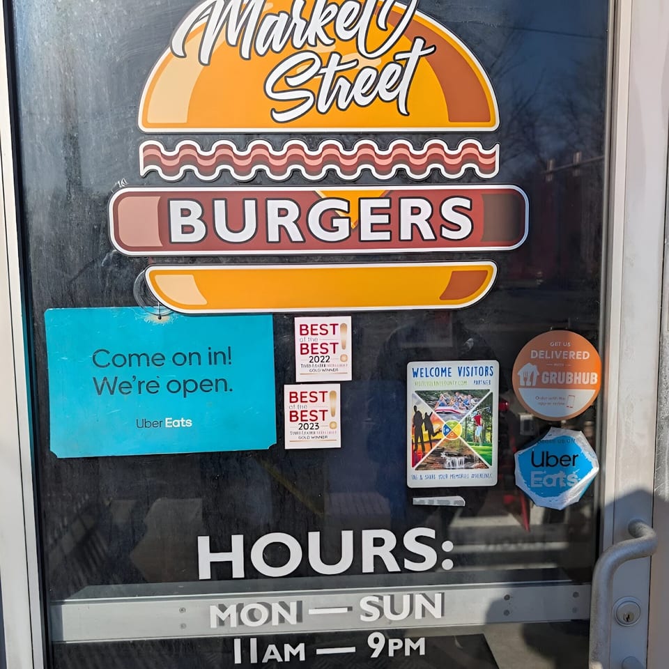 Market Street Burgers sign on a glass door with 'Open' and 'Hours' signs, plus delivery service stickers.